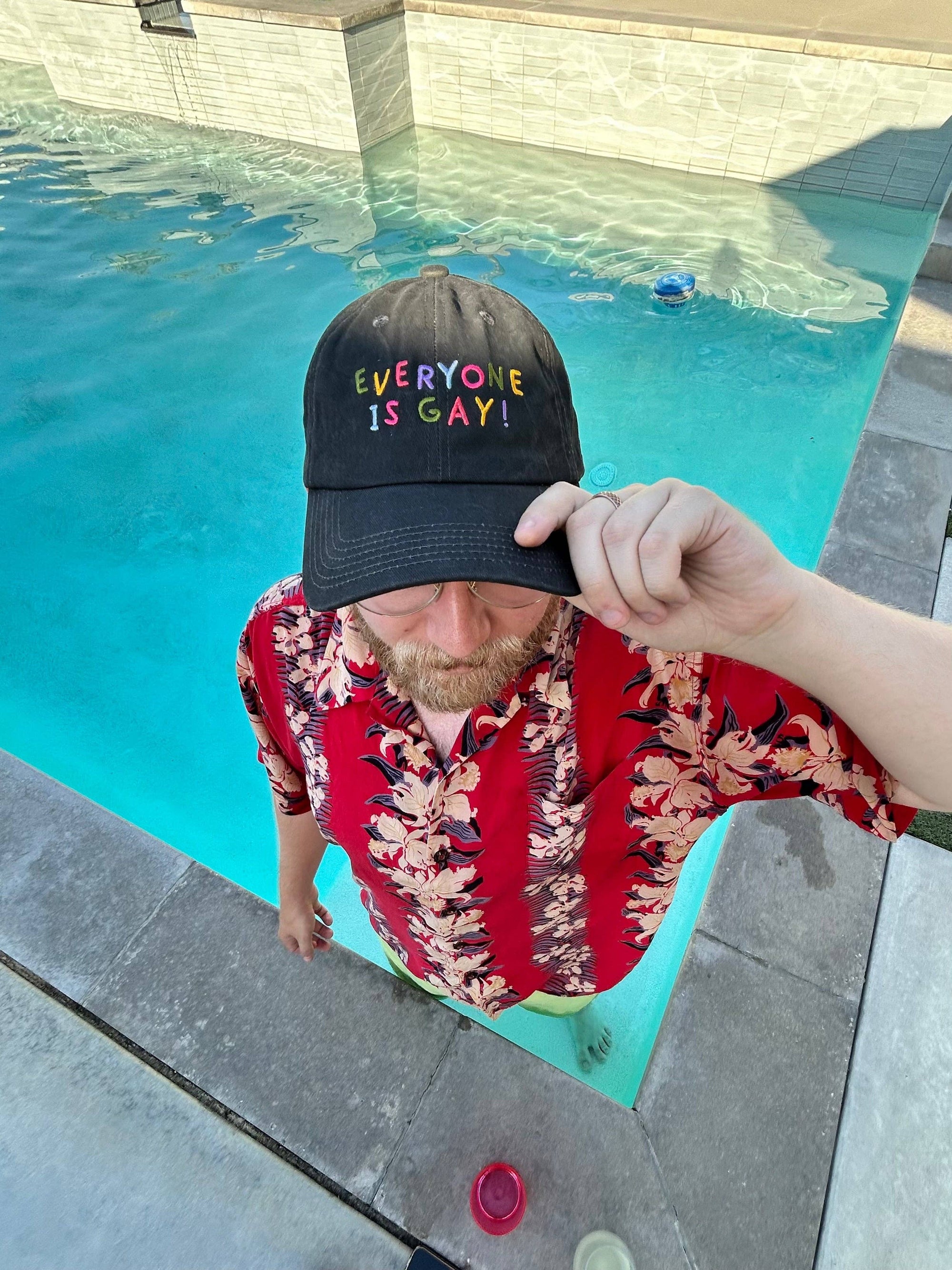 Person wearing a colorful 'Everyone is Gay!' cap by a pool