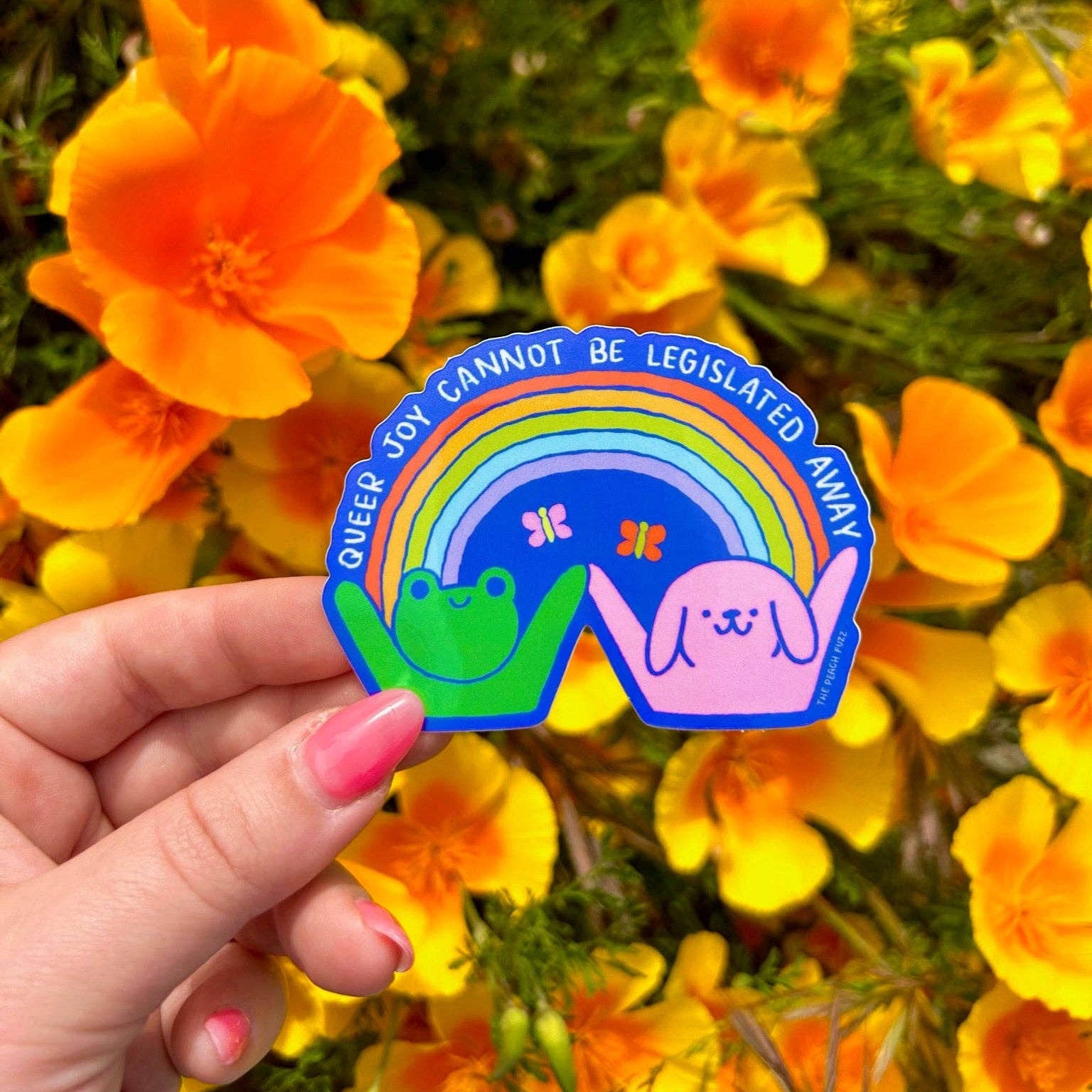Hand holding a colorful sticker with rainbow design and text in front of yellow flowers - Queer Joy Cannot Be Legislated Away 