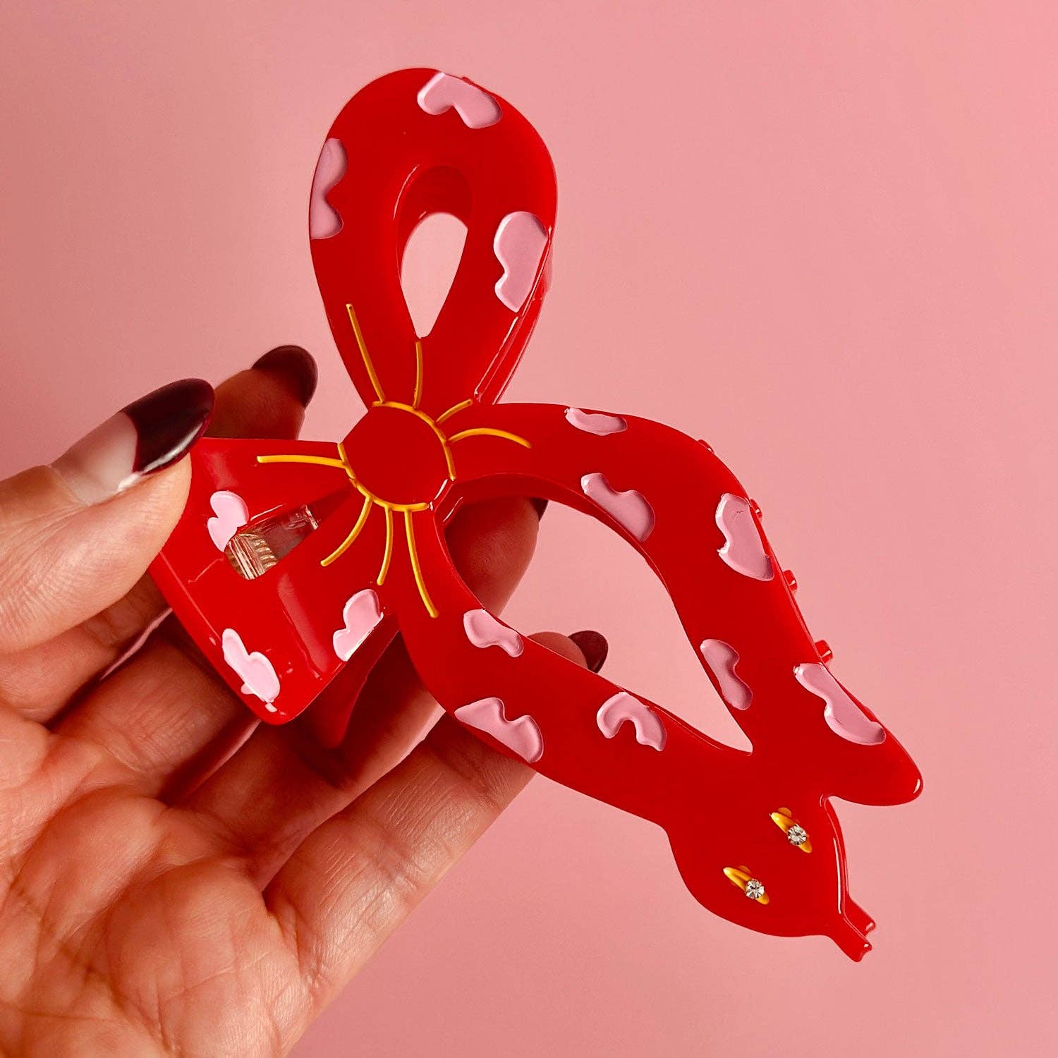 Red hair clip that is a ribbon with a snake face with bow design held by a hand against a pink background - year of the snake