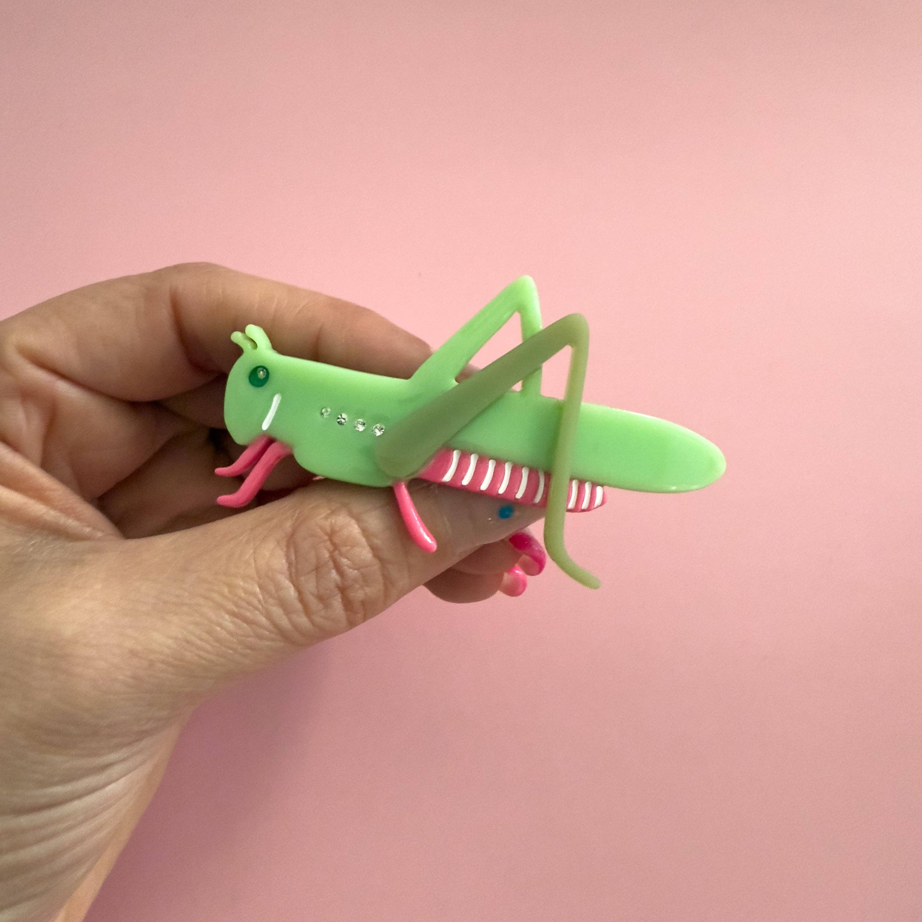 Hand holding a green and pink grasshopper hair clip against a pink background