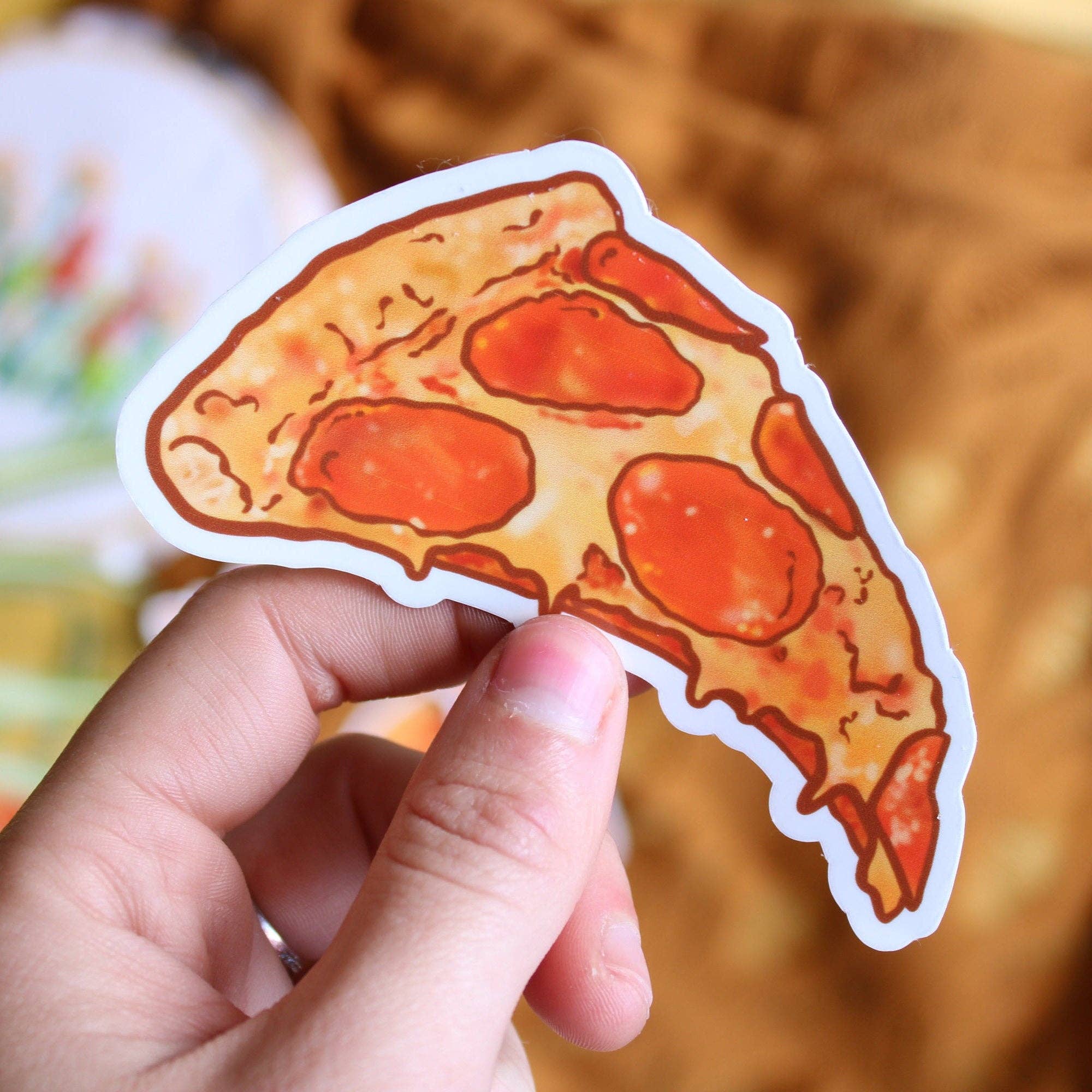 Hand holding a pepperoni pizza slice sticker against a blurred background