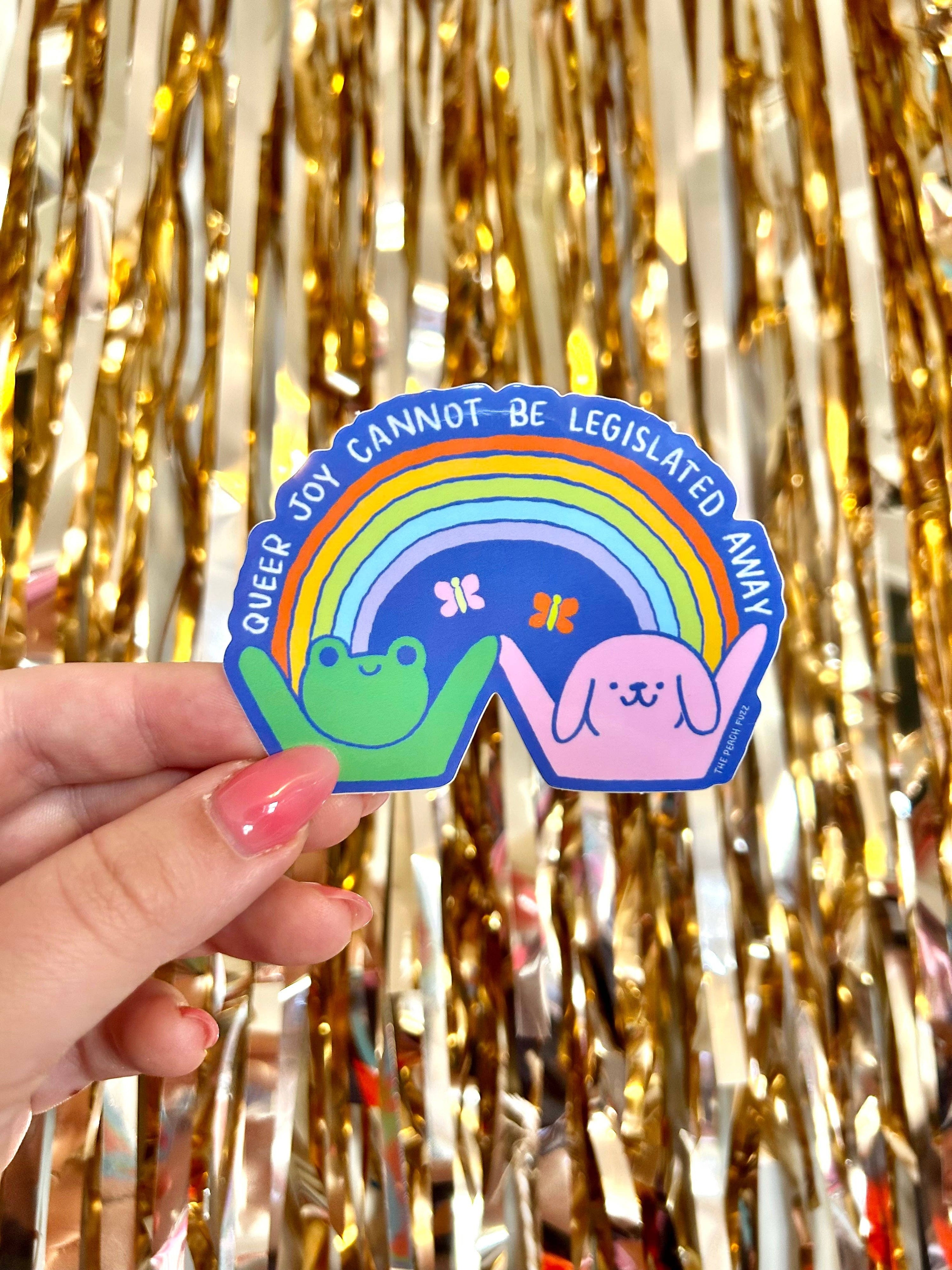 Sticker with a dog and frog and rainbow design held by a hand against a gold tinsel background - Queer Joy Cannot Be Legislated Away 