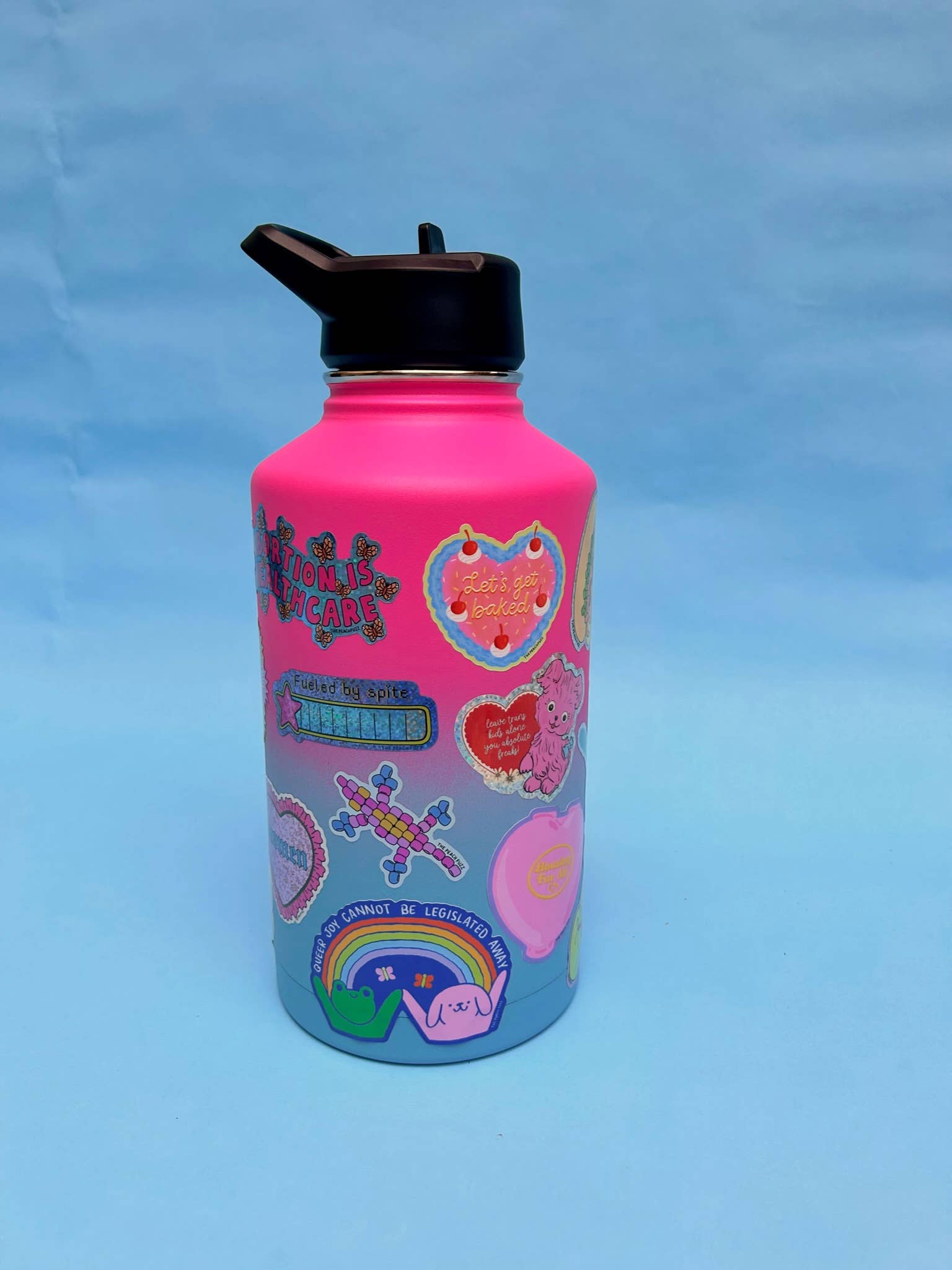 Pink water bottle with colorful designs on a blue background - Queer Joy Cannot Be Legislated Away 