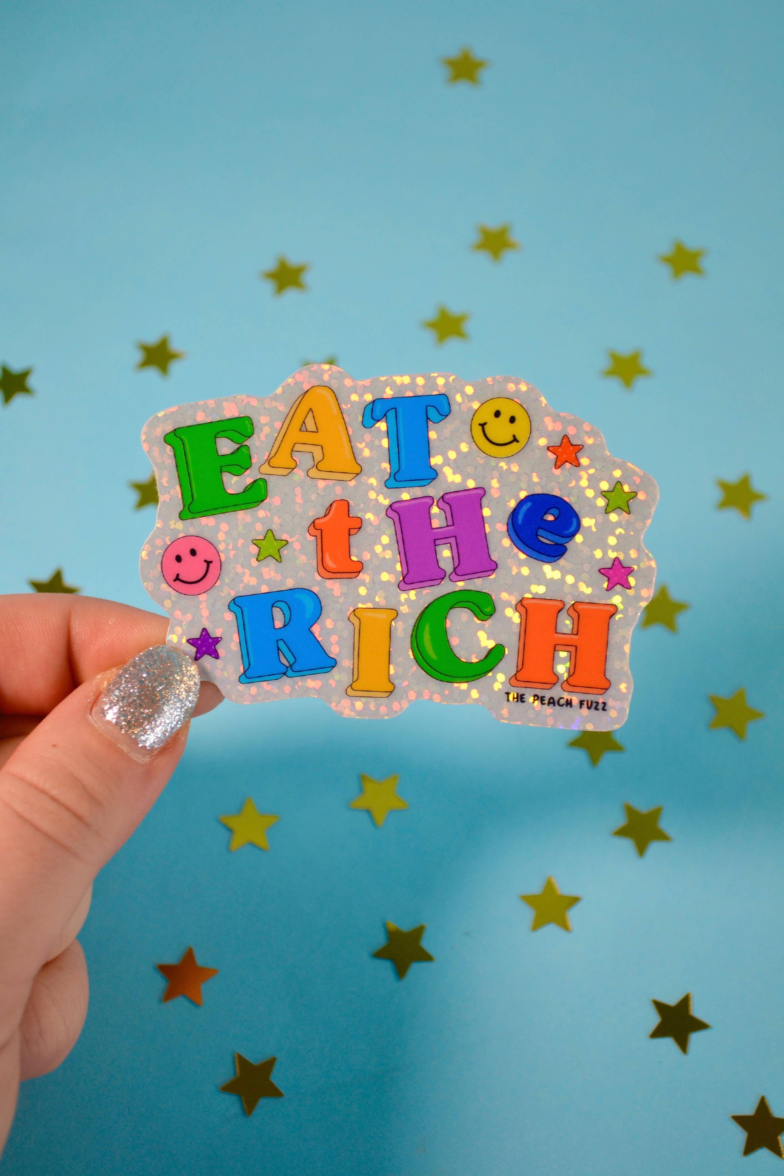 Colorful 'Eat the Rich' sticker held by a hand against a blue background with stars.