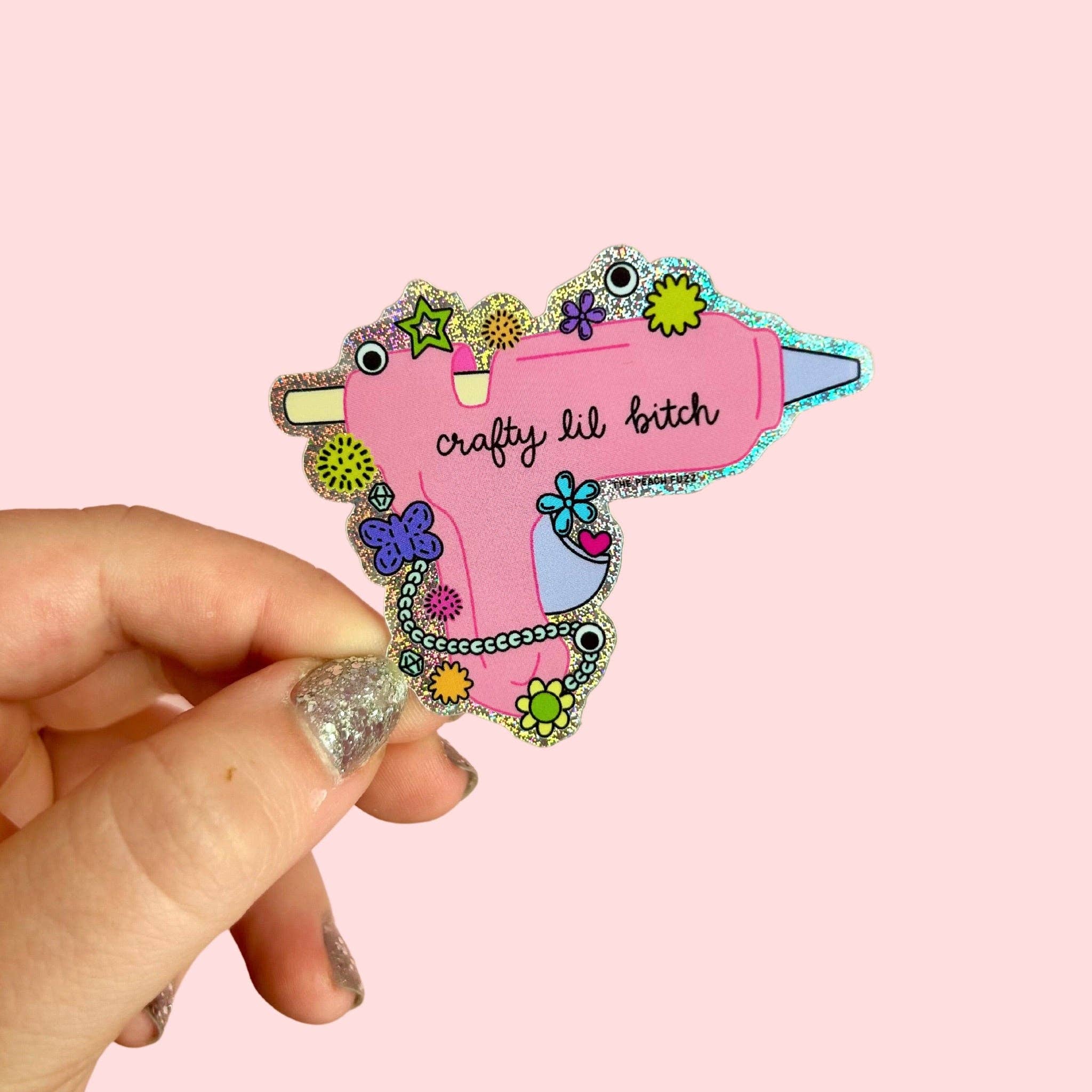 Colorful sticker of a hot glue gun - crafty lil bitch - with text held by a hand against a pink background