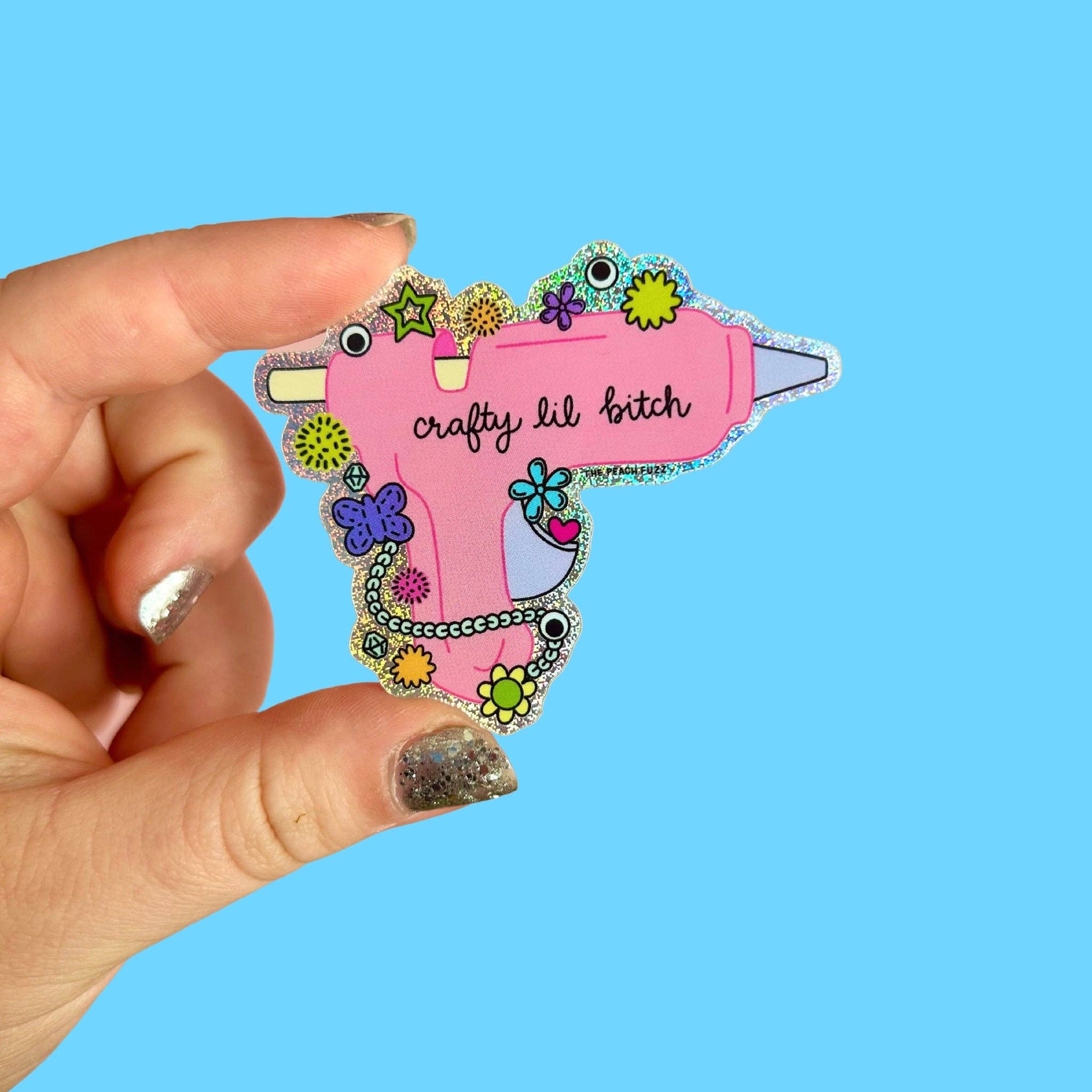 Hand holding a colorful hot glue gun -shaped glitter sticker with text against a blue background