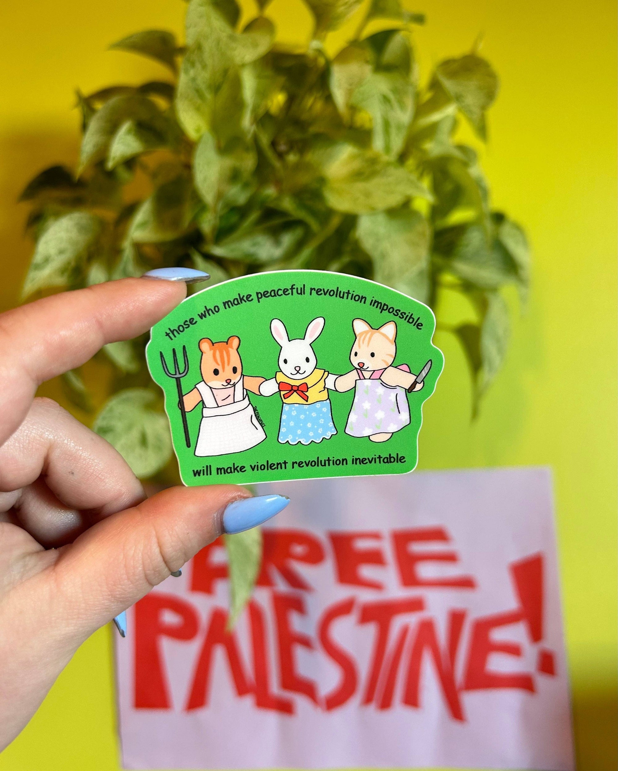 Hand holding a sticker with cartoon characters and text, with a 'Free Palestine' sign in the background.