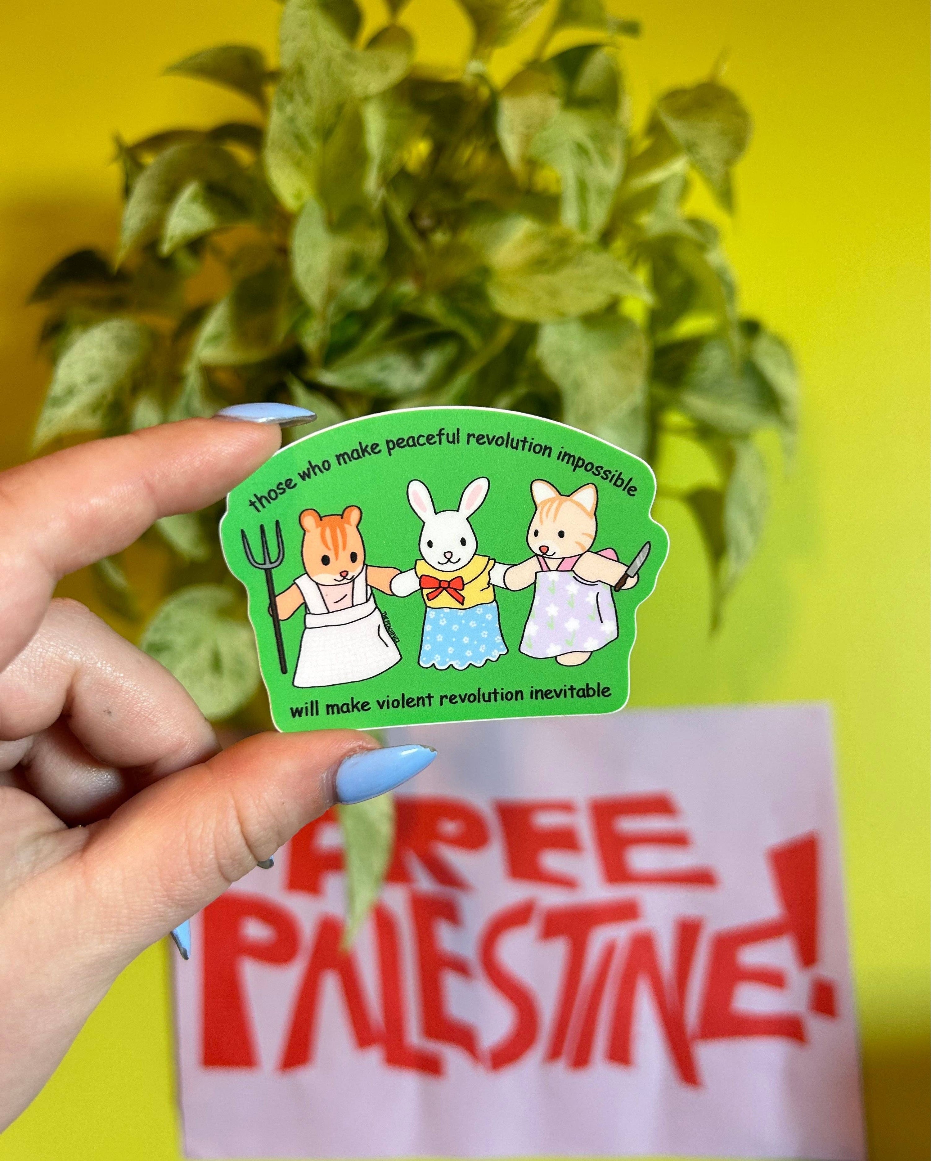 Hand holding a sticker with cartoon characters and text, with a 'Free Palestine' sign in the background.