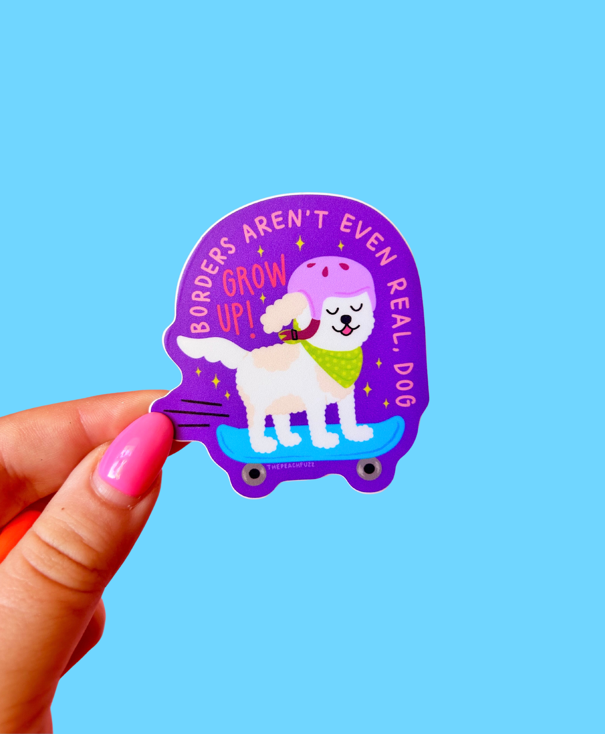 Sticker with a dog on a skateboard and text held by a hand against a blue background - borders aren't even real dog grow up