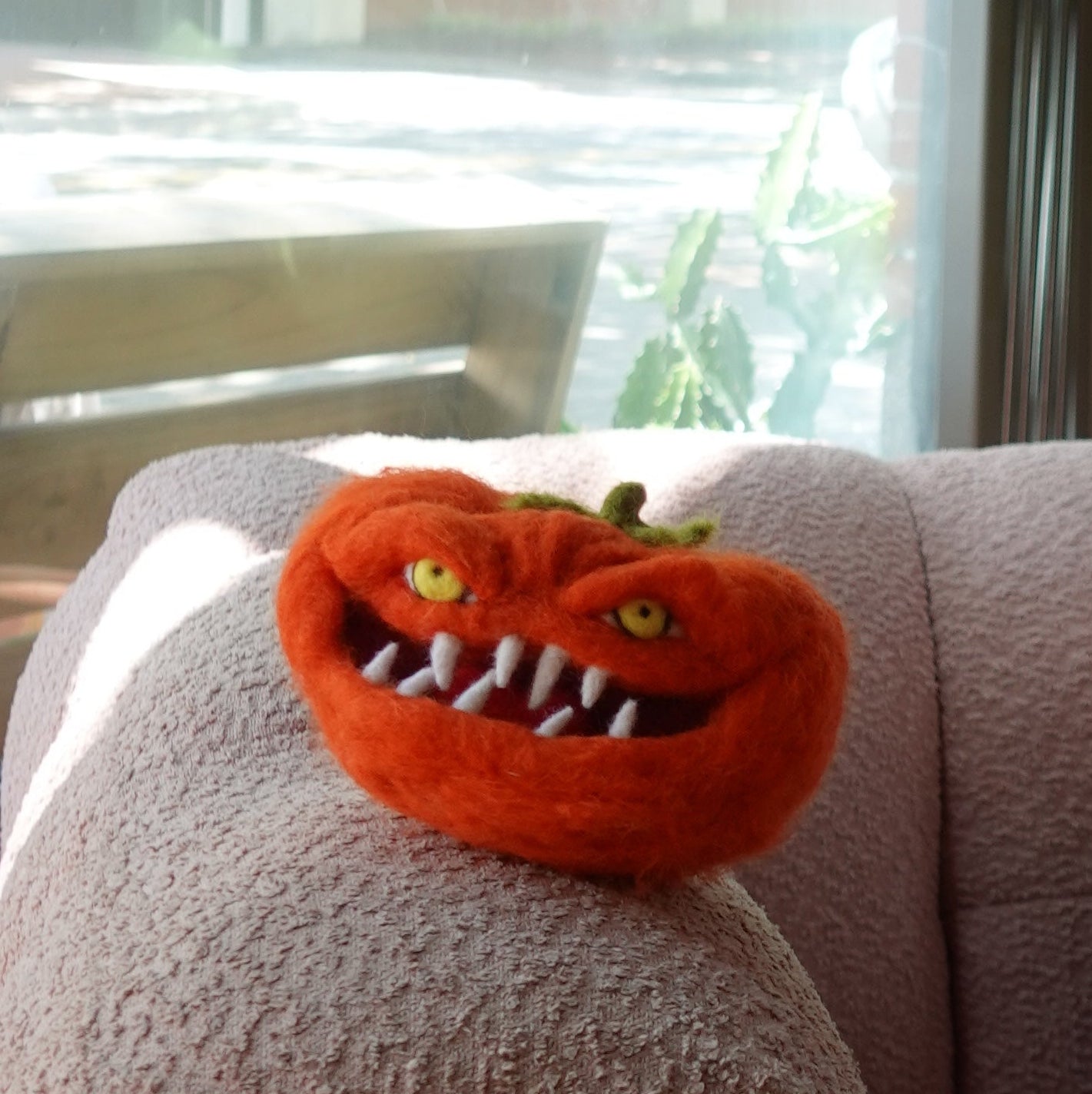 A small orange red felt tomato plush with a mischievous face, yellow eyes, and sharp white teeth, resting on the arm of a textured pink armchair near a window