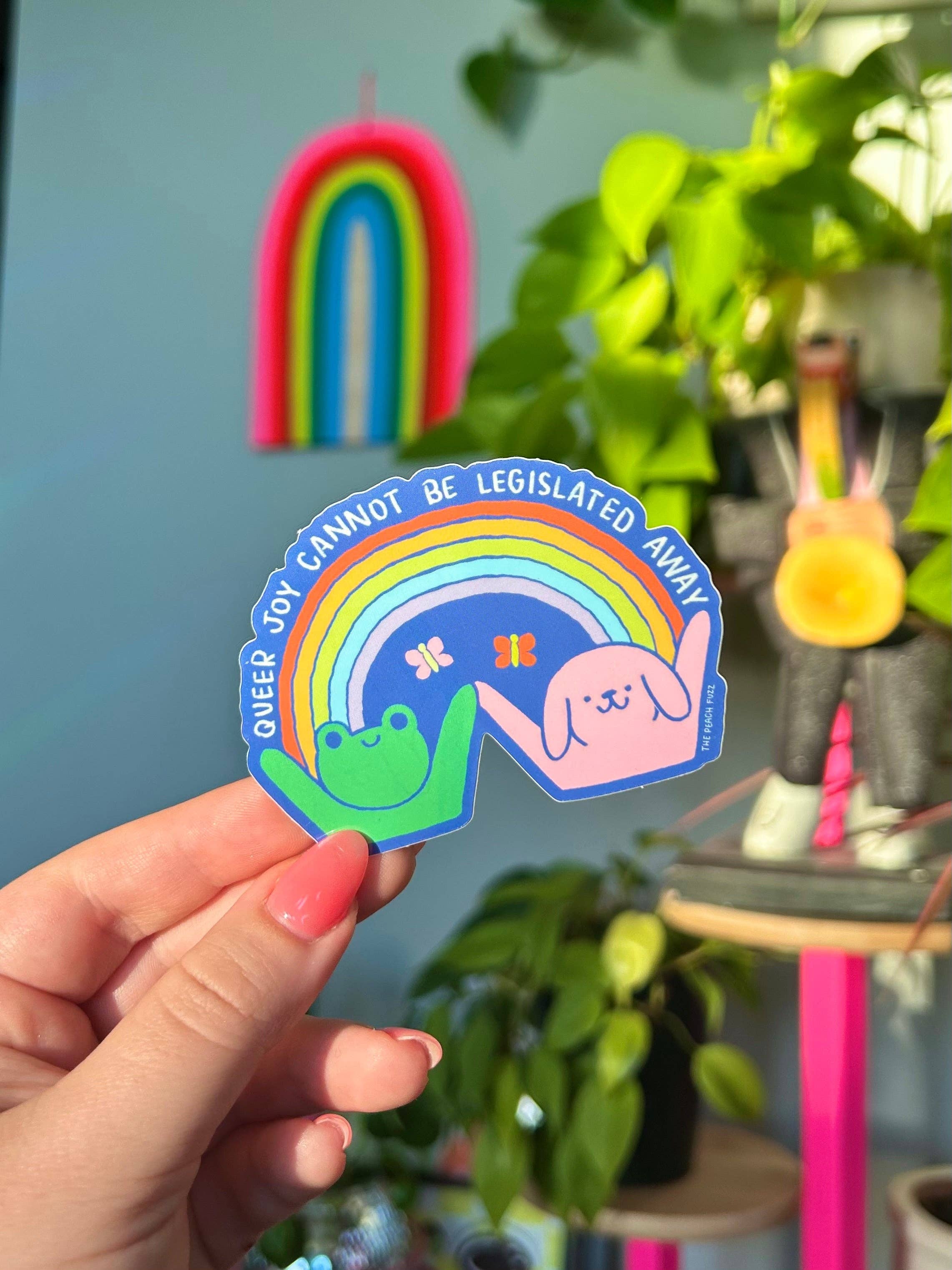 Hand holding a colorful sticker with a rainbow dog and frog and text, with plants and a colorful arch in the background. Queer Joy Cannot Be Legislated Away 