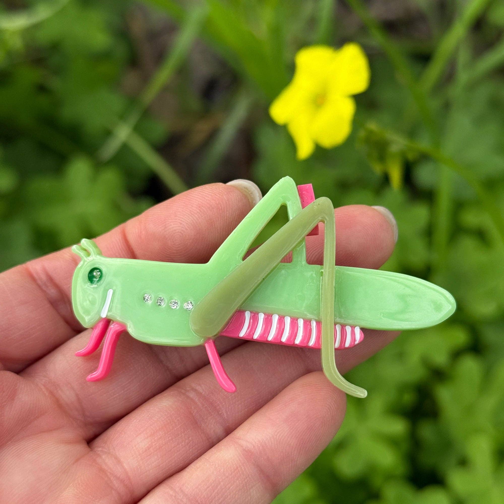 Green and pink grasshopper-shaped hair clip held in a hand with a blurred natural background
