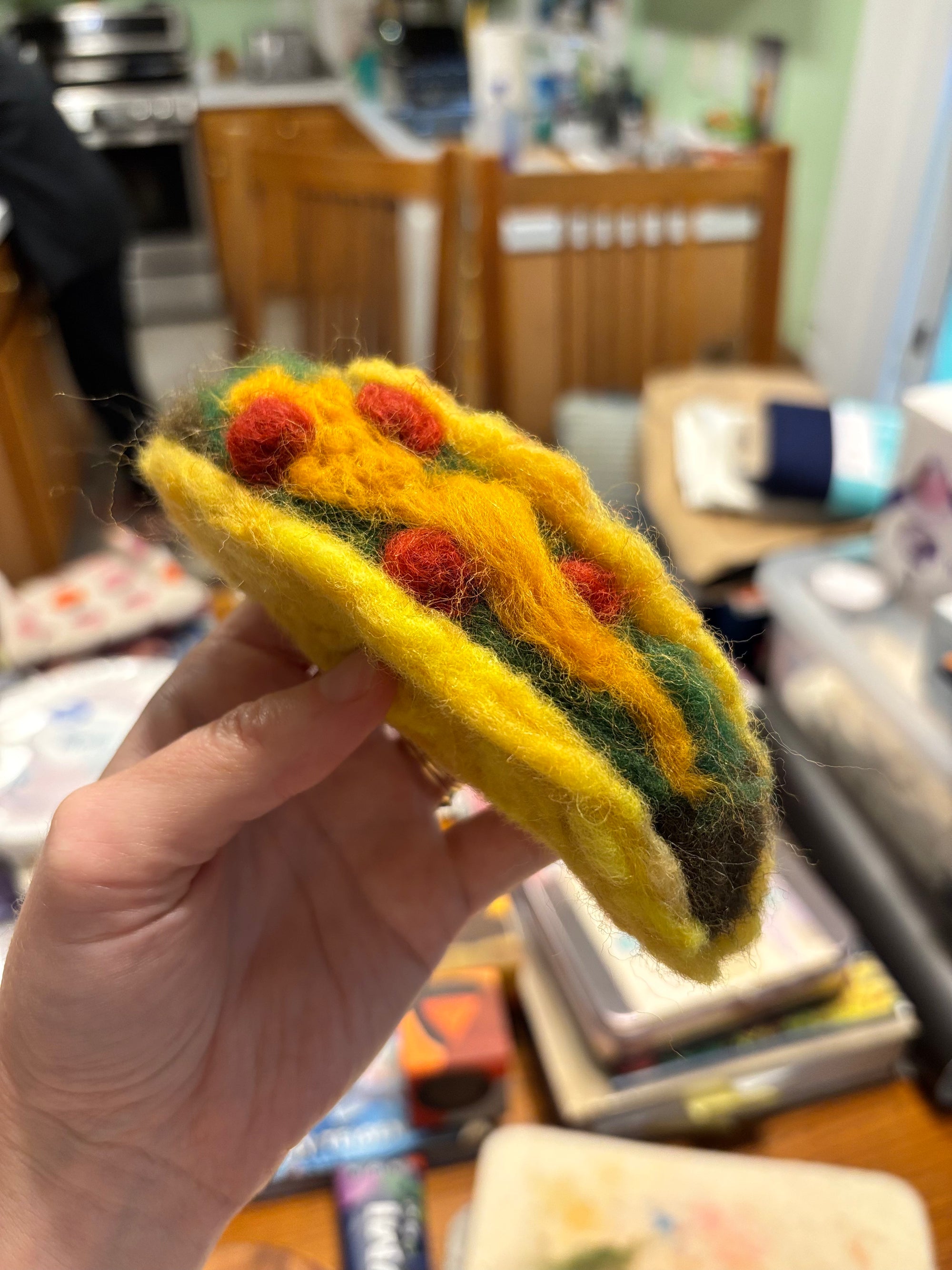 Hand holding a small felted taco craft with red and green details against a blurred indoor background