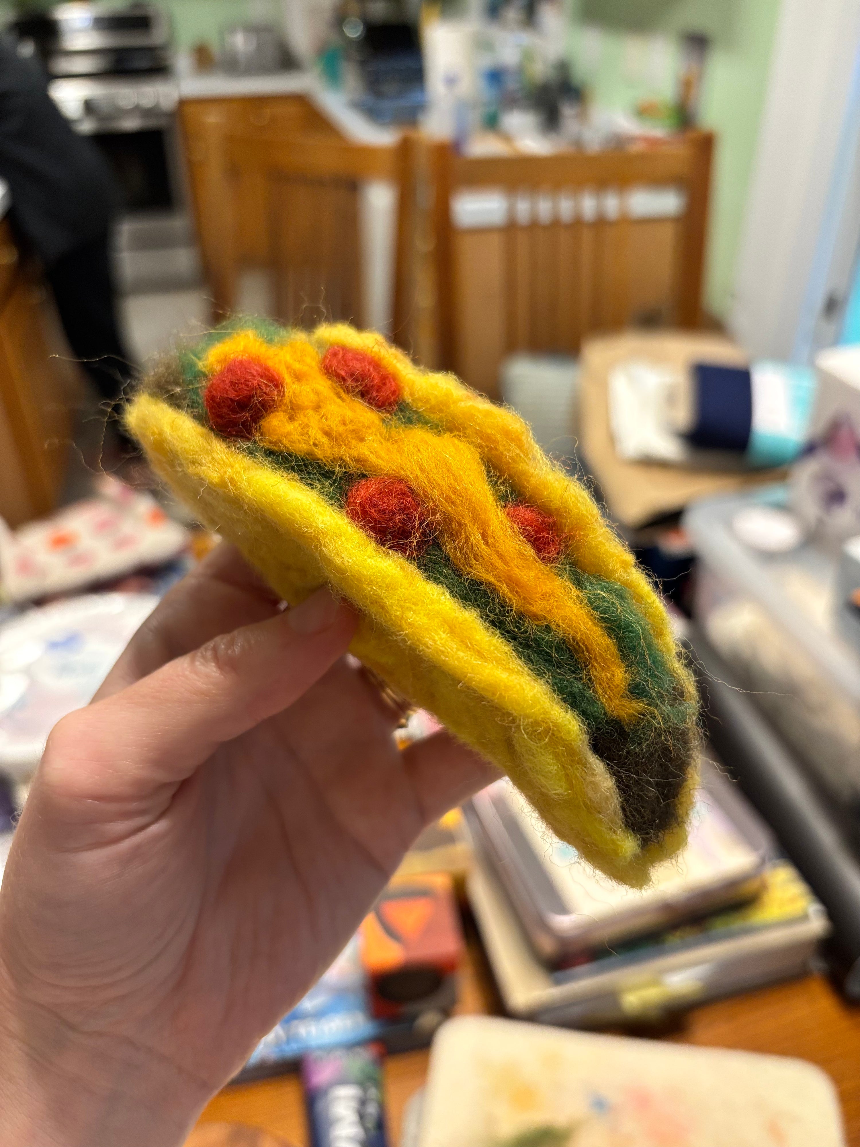 Hand holding a small felted taco craft with red and green details against a blurred indoor background