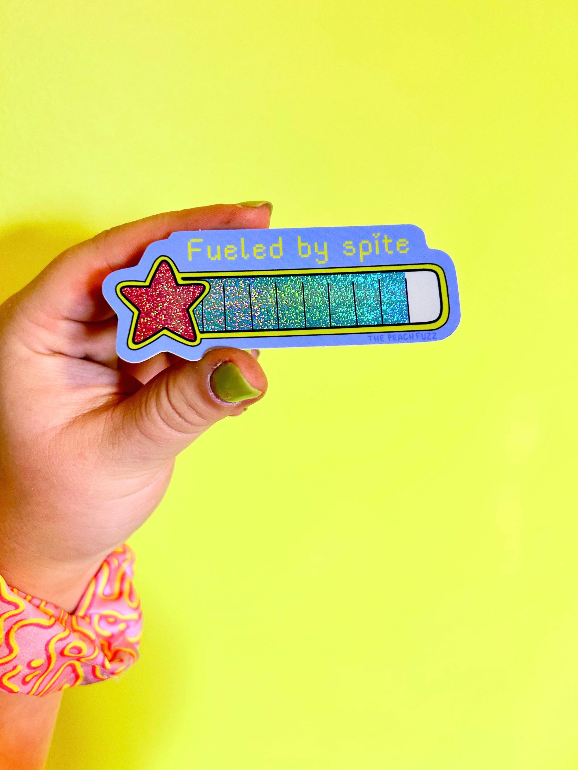 Hand holding a colorful battery-themed sticker with text against a yellow background