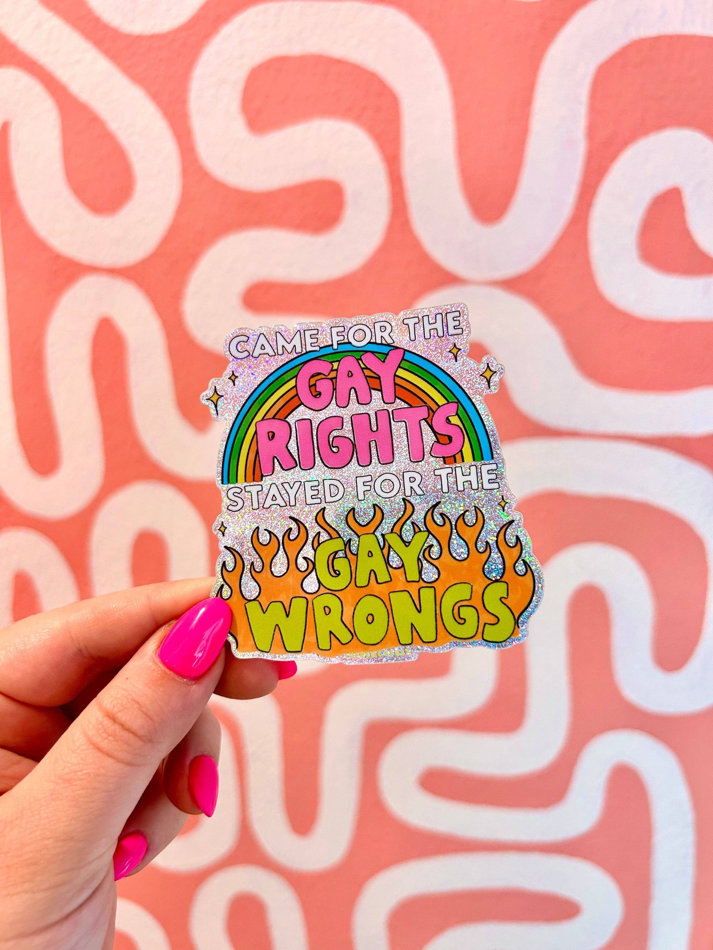 Sticker with rainbow design and text held by a hand against a pink background with white patterns