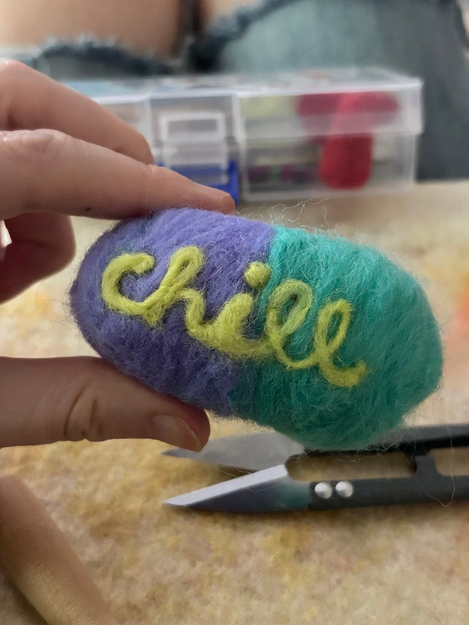 Multicolored needle felted pill with 'chill' text, held by a hand, with scissors and thread box in the background.
