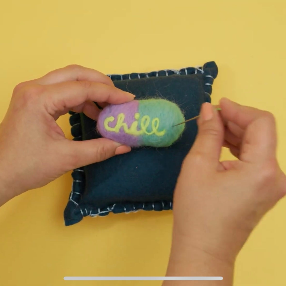 Person felting a colorful pill on a needle felting mat with 'chill' text onto a blue fabric background