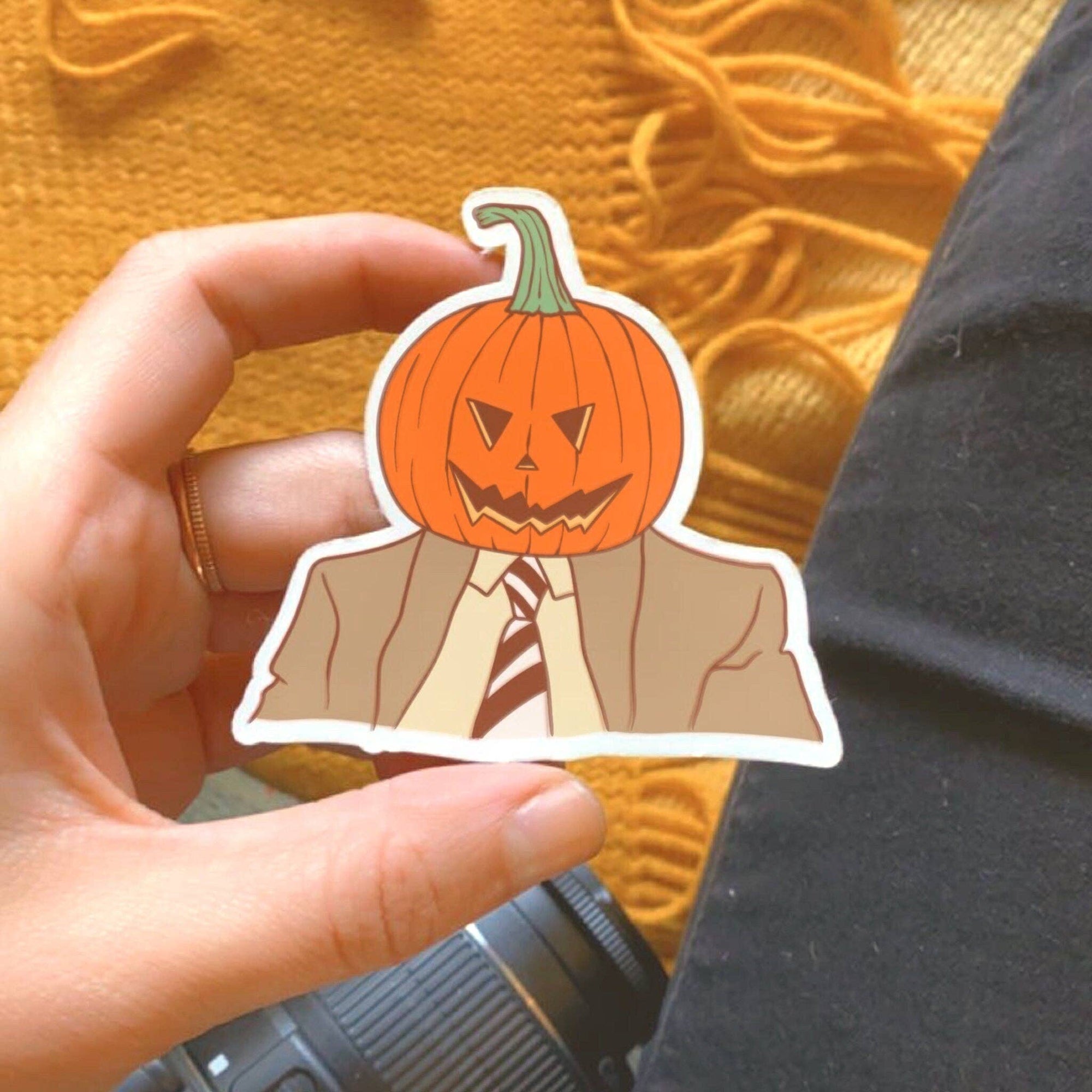 Hand holding a sticker of a person with a pumpkin head against an orange fabric background