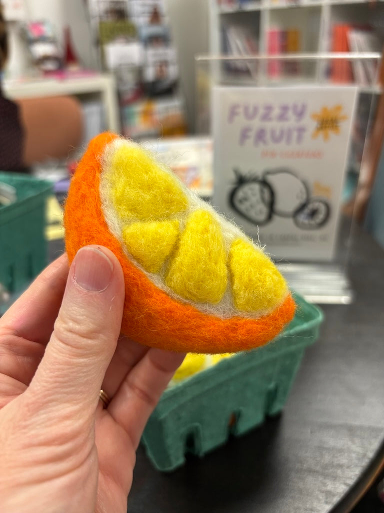 Hand holding a handmade felt orange slice pin cushion with a fuzzy fruit sign in the background