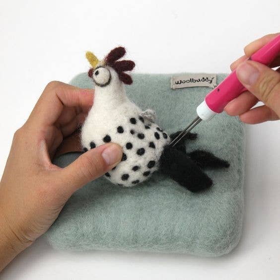 Small needle felted rooster toy on a light blue cushion with a pink tool being used.