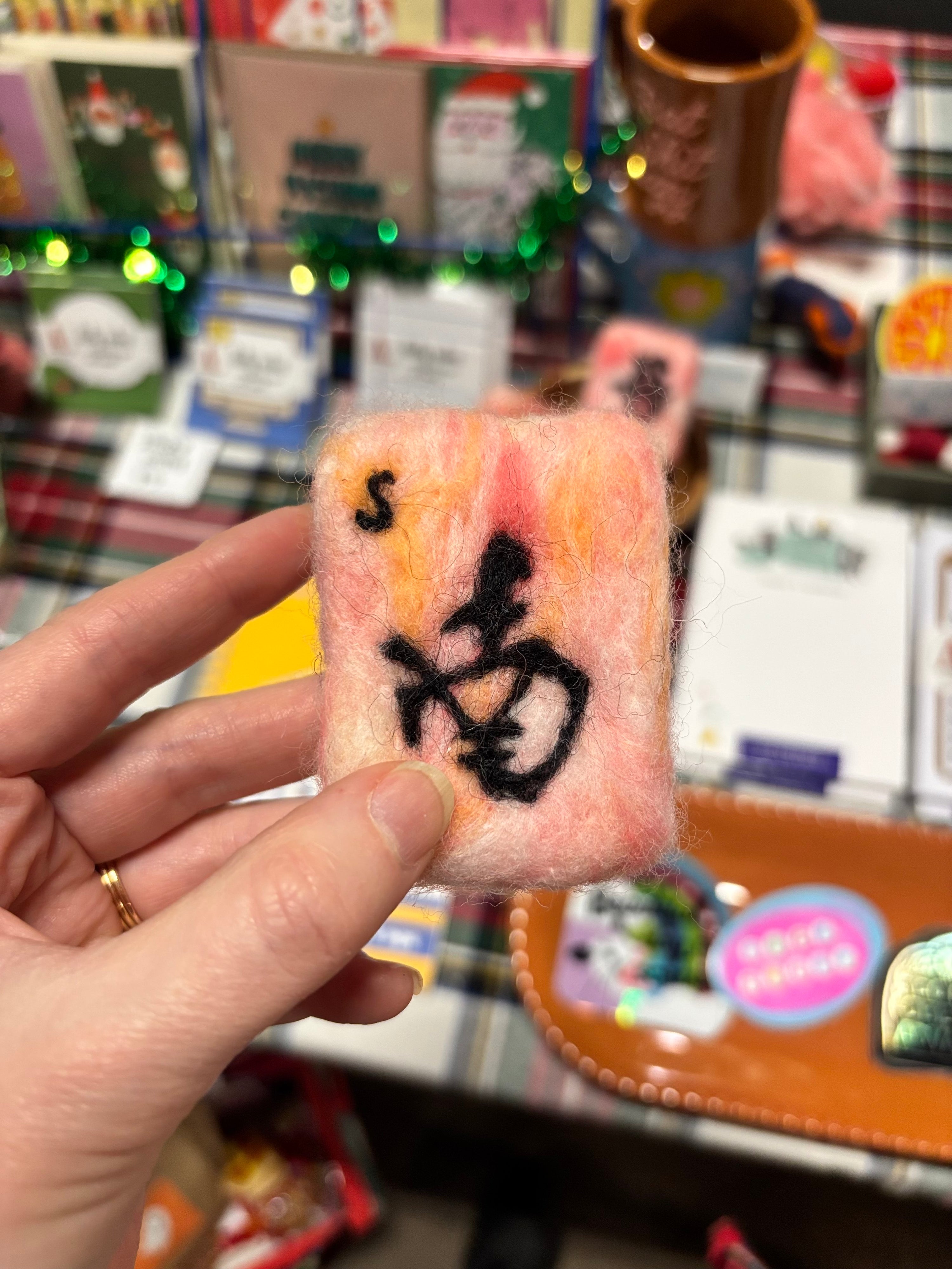 Hand holding a small felted pink and black textured mahjong wind tile product with a blurred store background needle felting