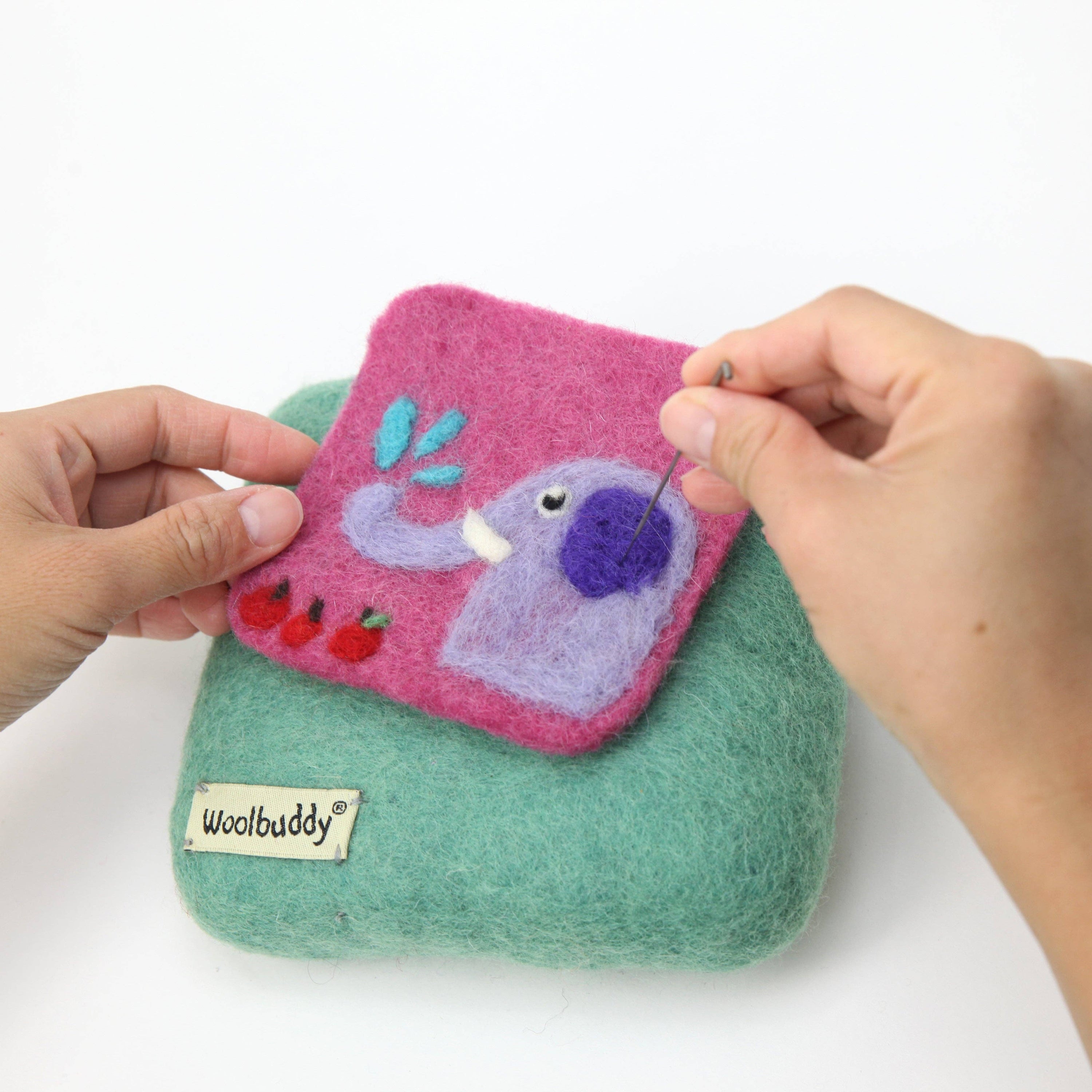 Handmade felted on a teal woolbuddy felting mat with a purple  and pink elephant creature design, held by hands with a white background.