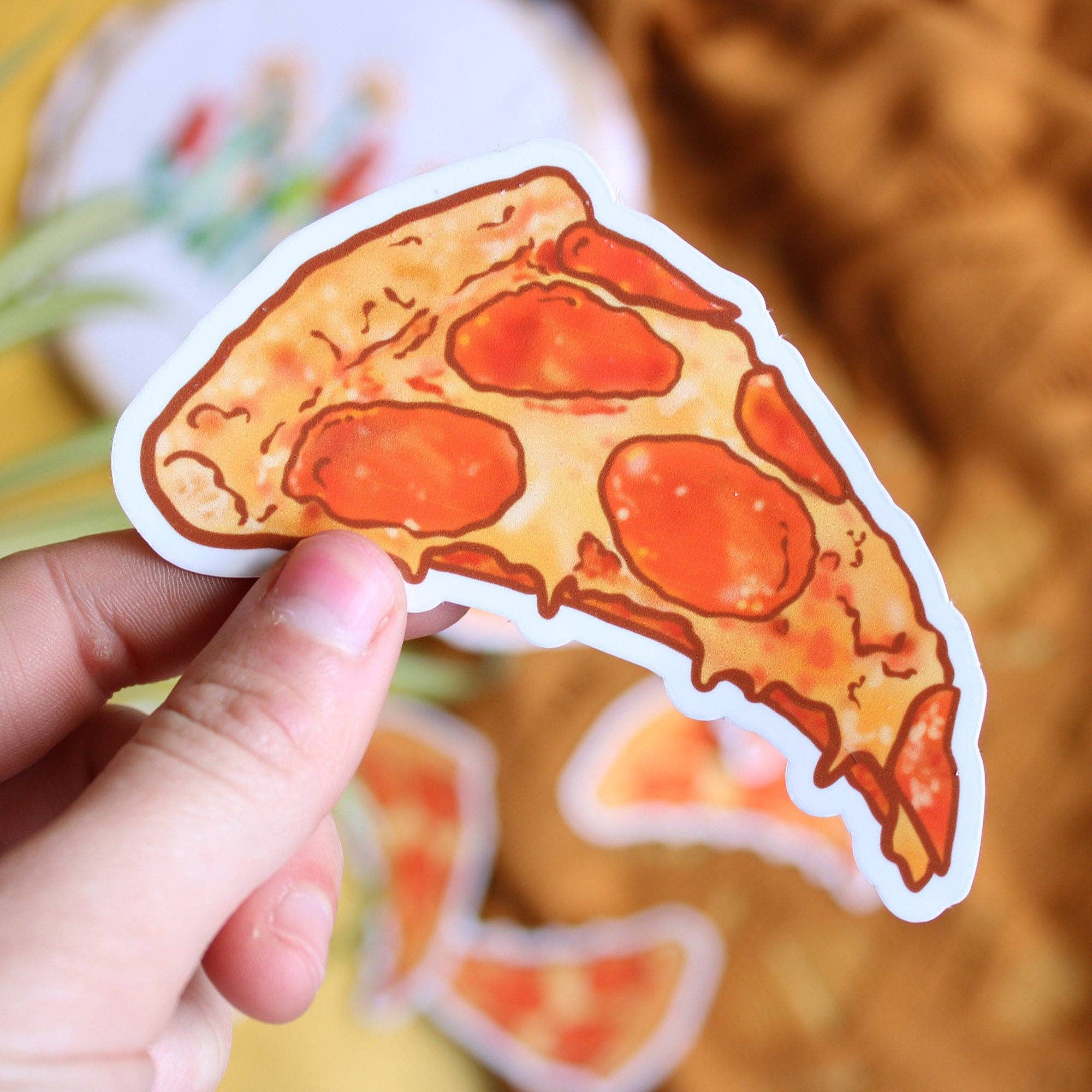 Hand holding a pepperoni pizza slice sticker with a blurred background