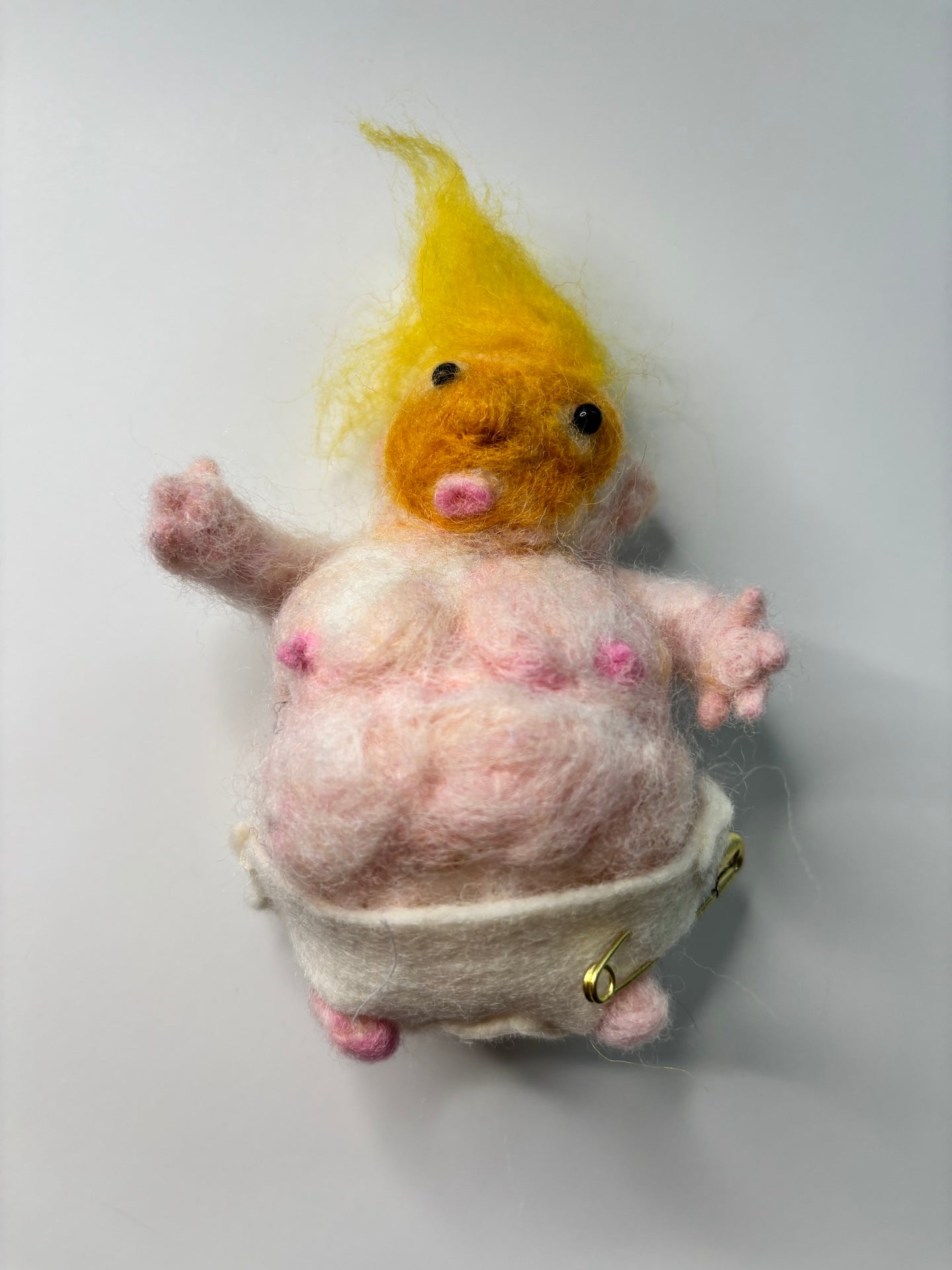 Small needle feltied plush toy of the ugly president with yellow hair, orange face, white diaper, and a pale pink body on a light gray background