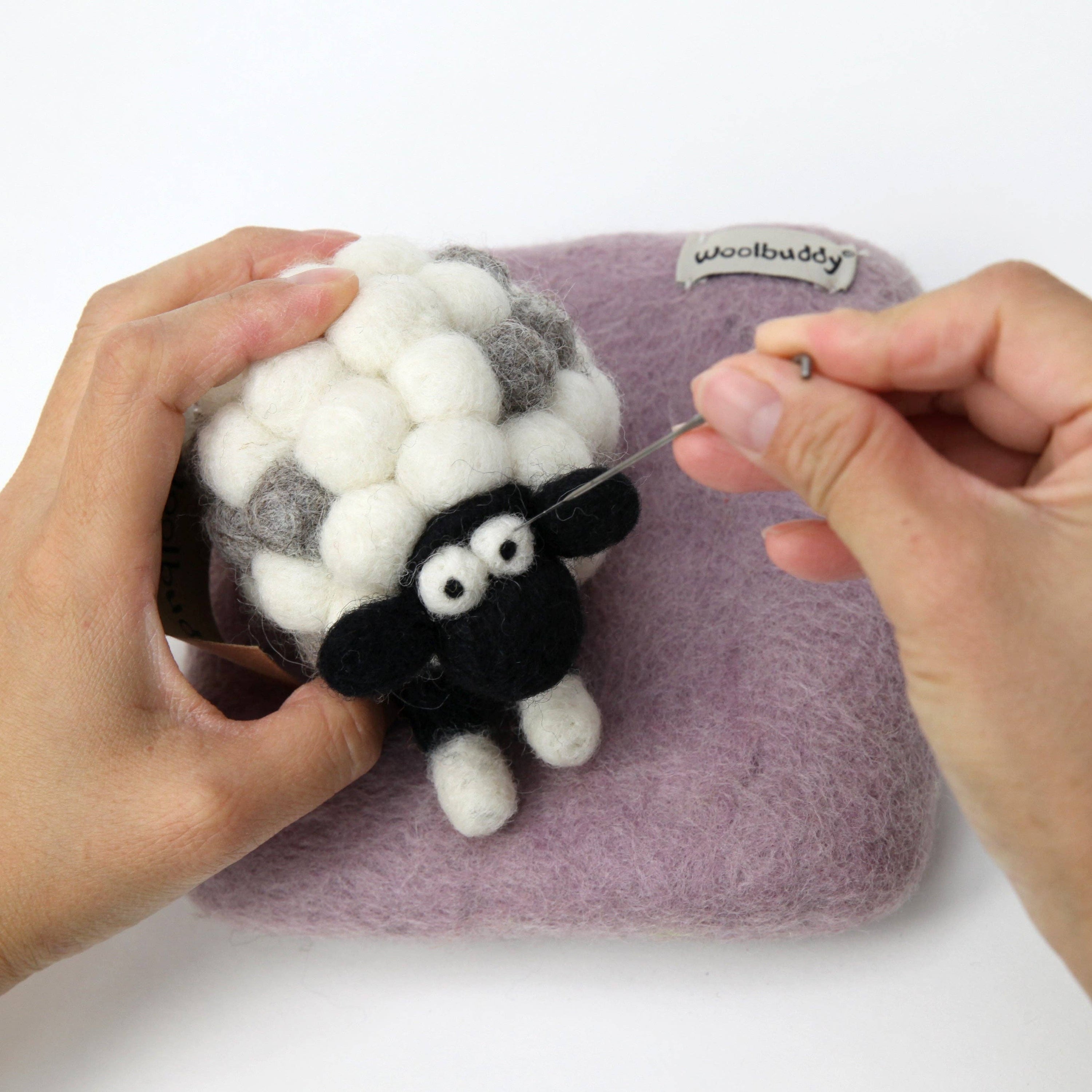 Person using a needle to sew a black and white sheep onto a purple fabric with 'woolbuddy' branding.